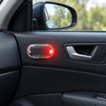 A small oval solar-powered warning light with a red flashing LED, mounted on a black car door panel by the interior handle.